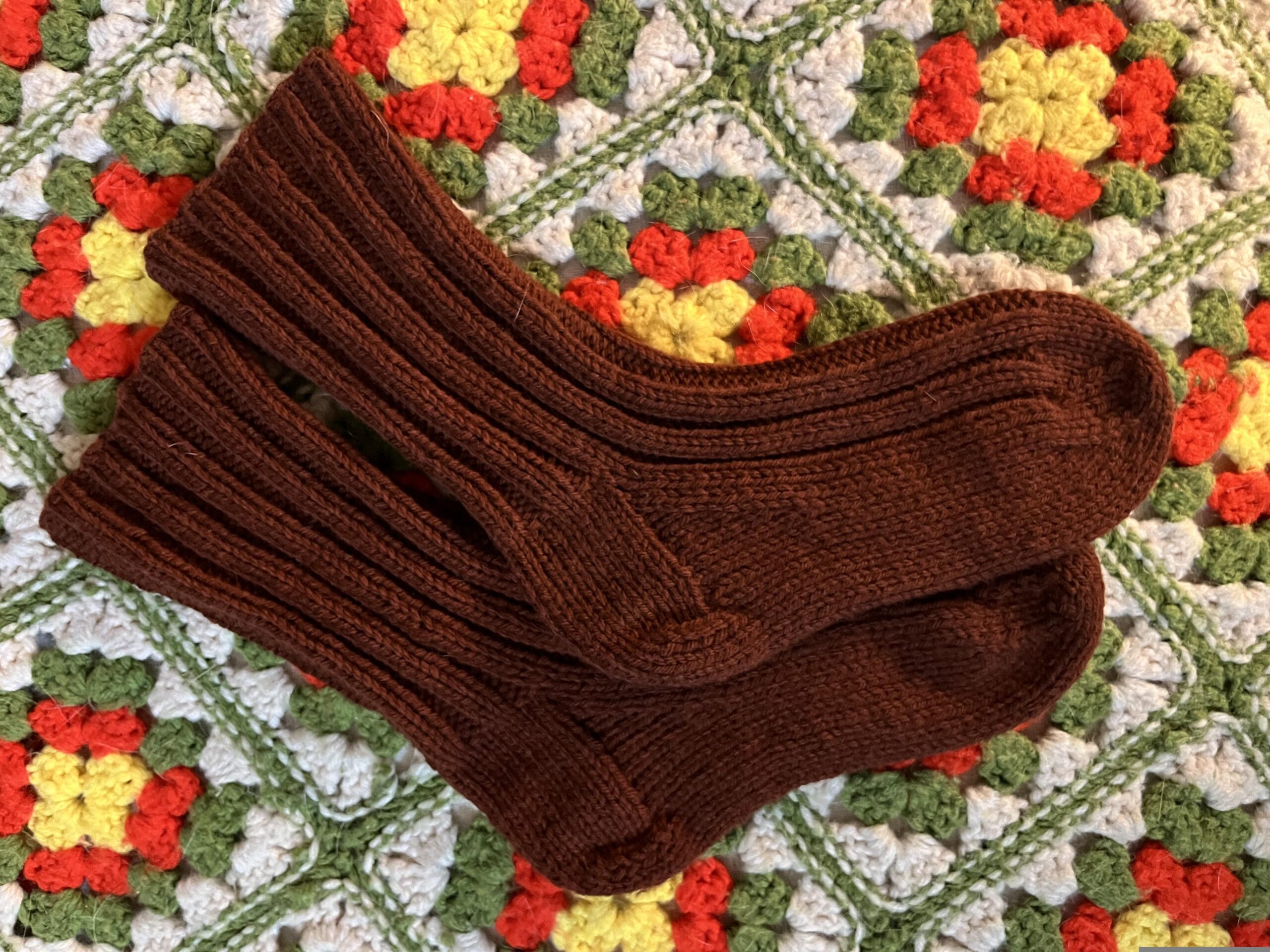 pair of hand-knit brown worsted weight socks on a vintage crochet granny square blanket in cream, avocado green, bright orange, and yellow.
